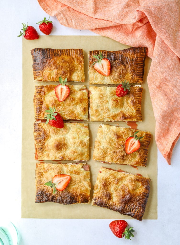 a strawberry rhubarb puff pastry tart cut into 8 rectangles on a blue background with fresh strawberries on top and a pink cloth napkin