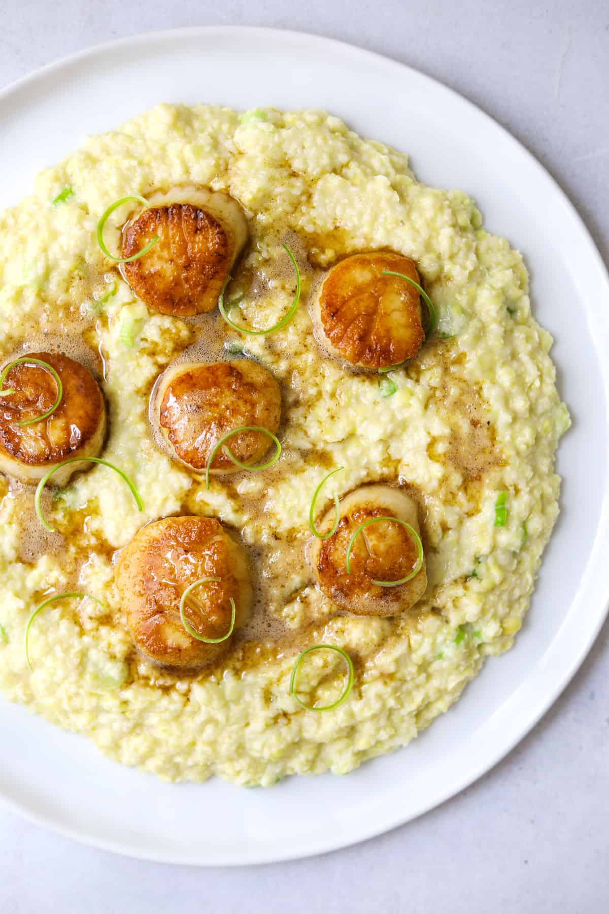 a plate of creamy corn porridge topped with golden-brown seared scallops, melted brown butter and sliced green onion