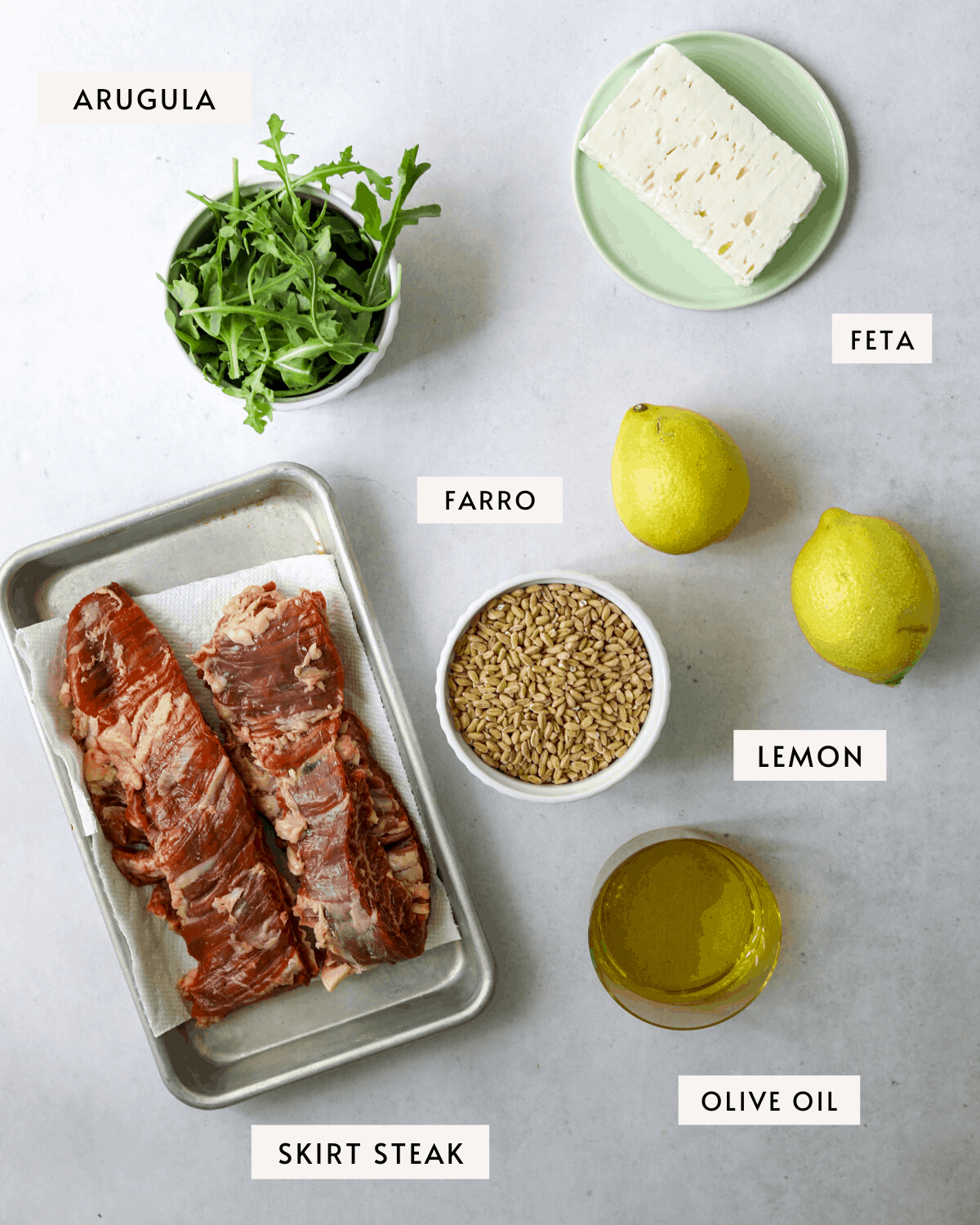recipe ingredients; a bowl of arugula, a tray of skirt steak, lemons, feta cheese, farro, olive oil.