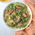 a white bowl filled with farro, arugula, feta cheese, and grilled steak on a blue background with a pink cloth napkin