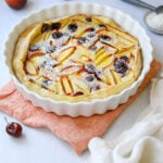 a white fluted baking dish filled with stone fruit clafoutis dusted in powdered sugar, surrounded by peaches, cherries and a scoop of vanilla ice cream