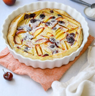 a white fluted baking dish filled with stone fruit clafoutis dusted in powdered sugar, surrounded by peaches, cherries and a scoop of vanilla ice cream