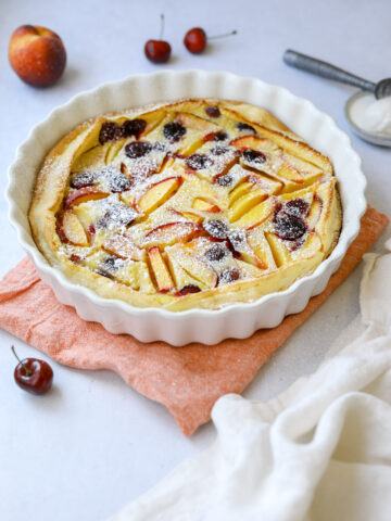 a white fluted baking dish filled with stone fruit clafoutis dusted in powdered sugar, surrounded by peaches, cherries and a scoop of vanilla ice cream
