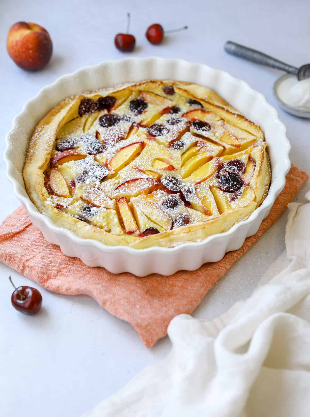 a white fluted baking dish filled with stone fruit clafoutis dusted in powdered sugar, surrounded by peaches, cherries and a scoop of vanilla ice cream 