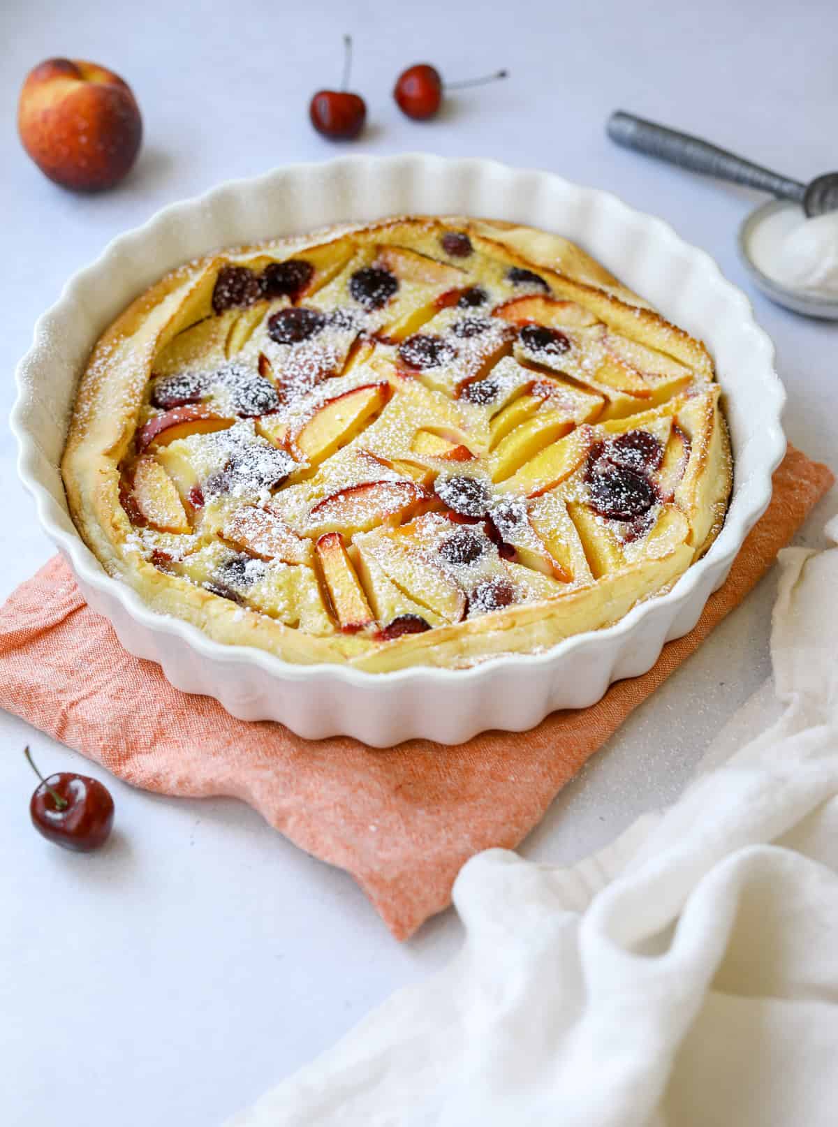 a white fluted baking dish filled with stone fruit clafoutis dusted in powdered sugar, surrounded by peaches, cherries and a scoop of vanilla ice cream