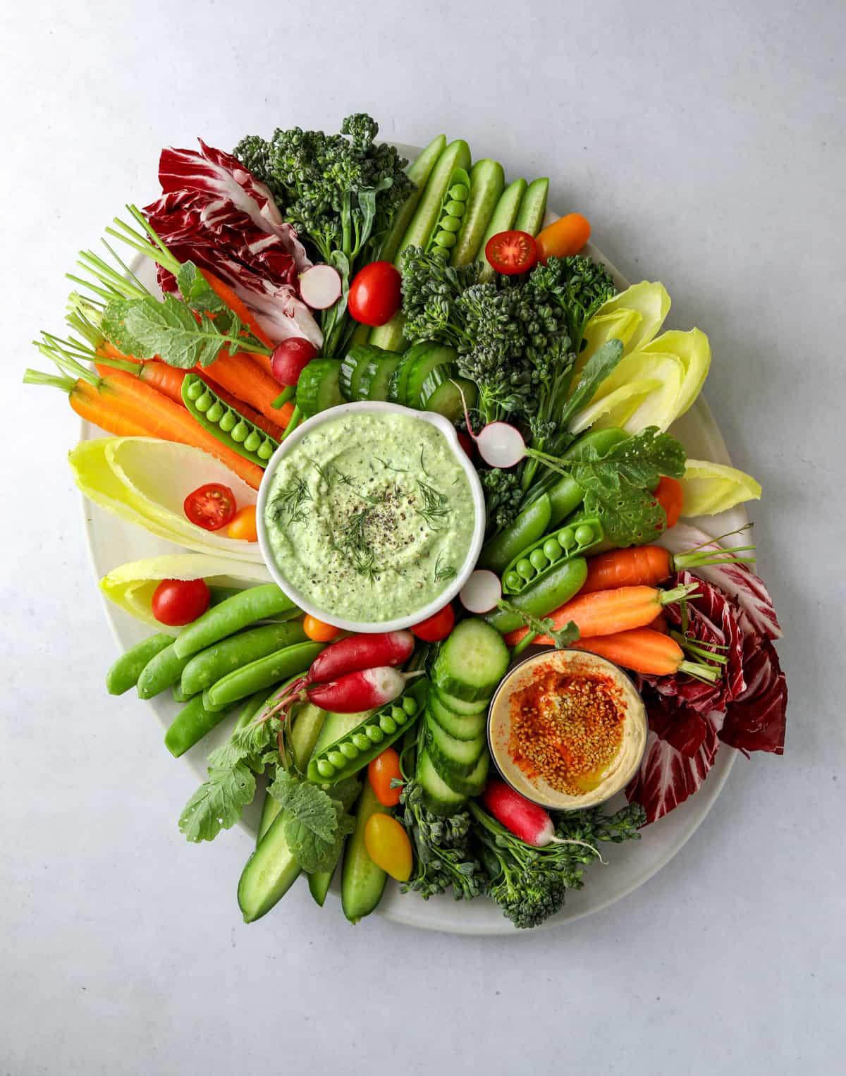 a vibrant an colorful array of vegetables (cucumbers, carrots, broccoli, radish) on an oval platter with two bowls of dip, one feta dip and one hummus.