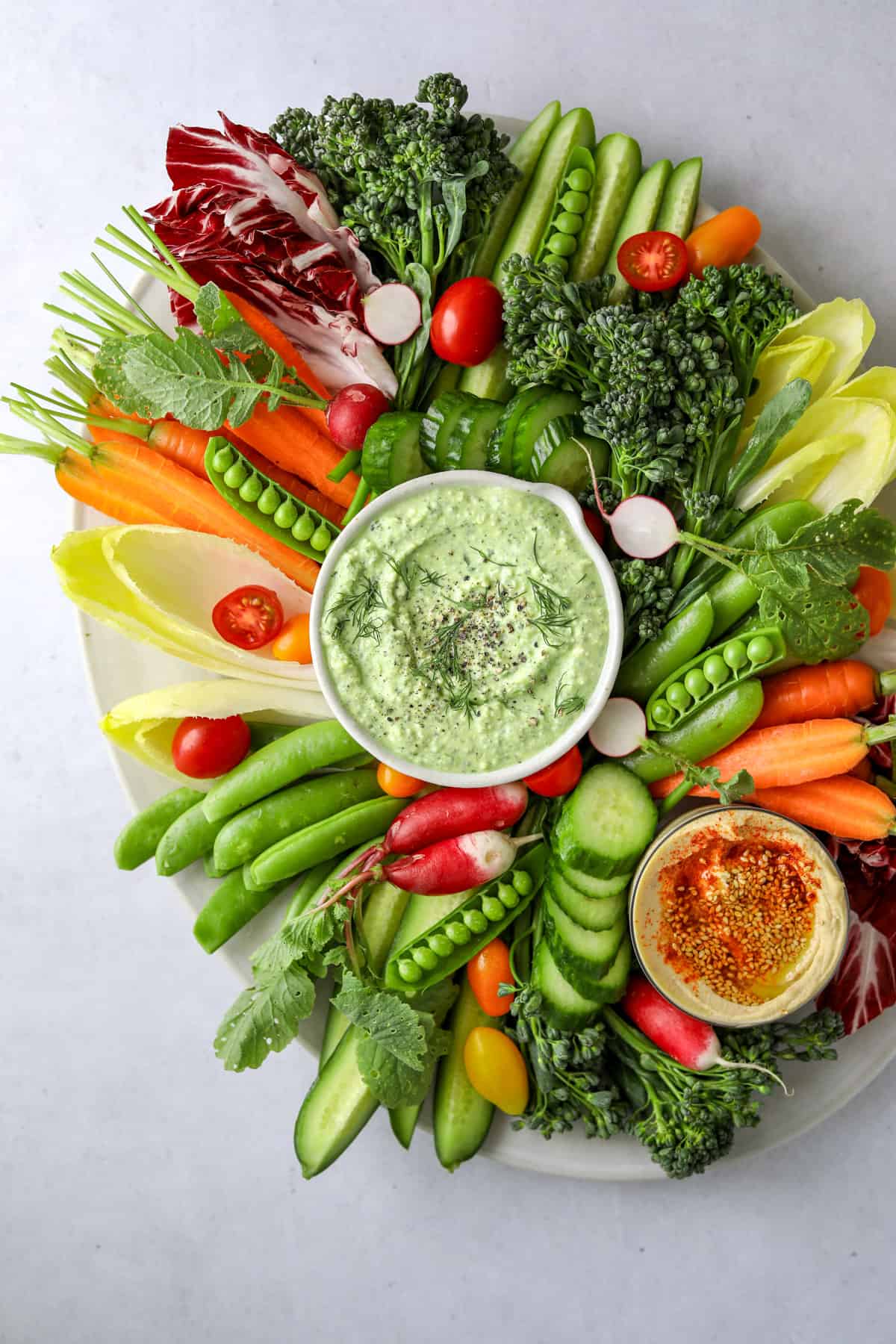 an oval crudités platter filled with cut vegetables like snap peas, carrots, broccolini, cucumber and two bowls of dips; feta dip and hummus