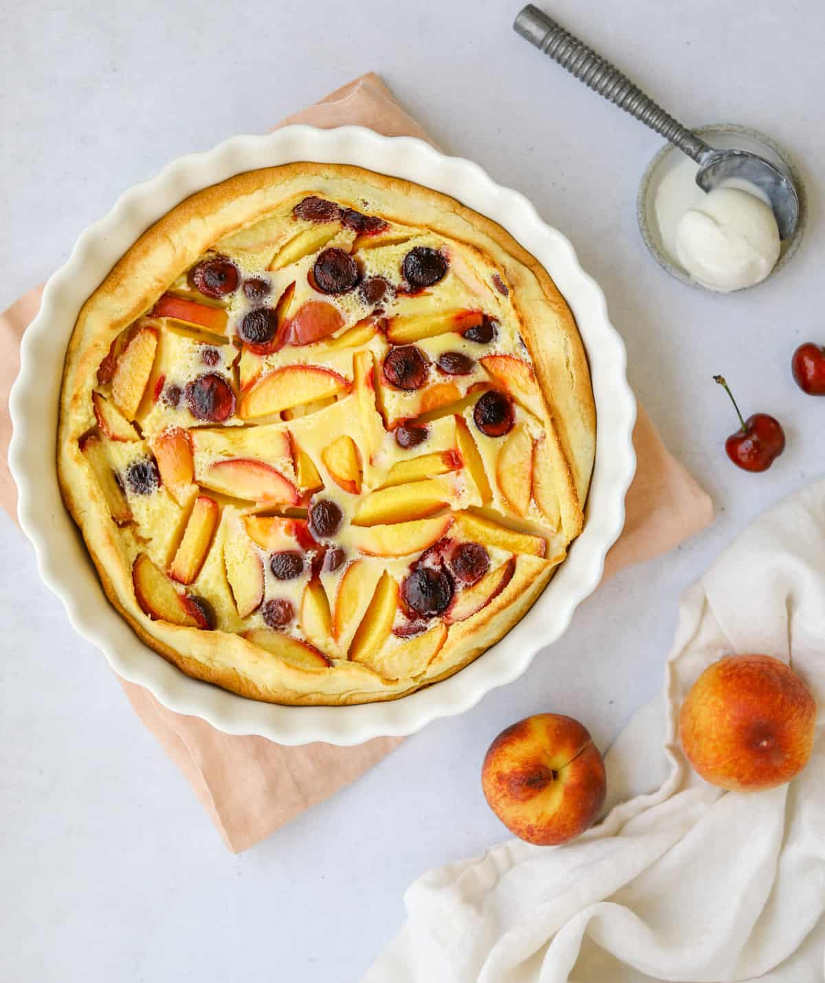 stone fruit clafoutis in a round white fluted baking dish on top of a pink linen napkin. a scoop of ice cream on the side.