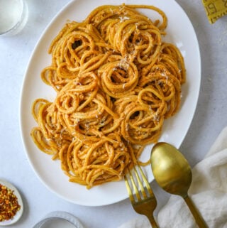 a white oval platter fulled with bucatini with sun-dried tomato pesto with gold serving utensils.