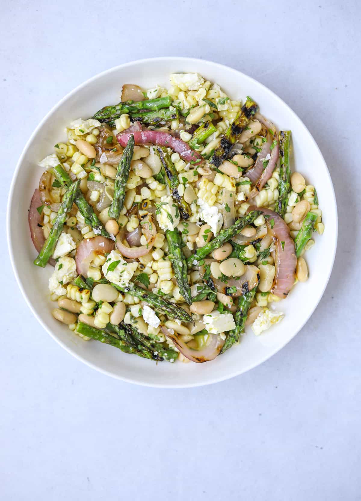 a white bowl filled with grilled vegetable salad with white beans, get feta cheese, asparagus, red onion, on a blue background