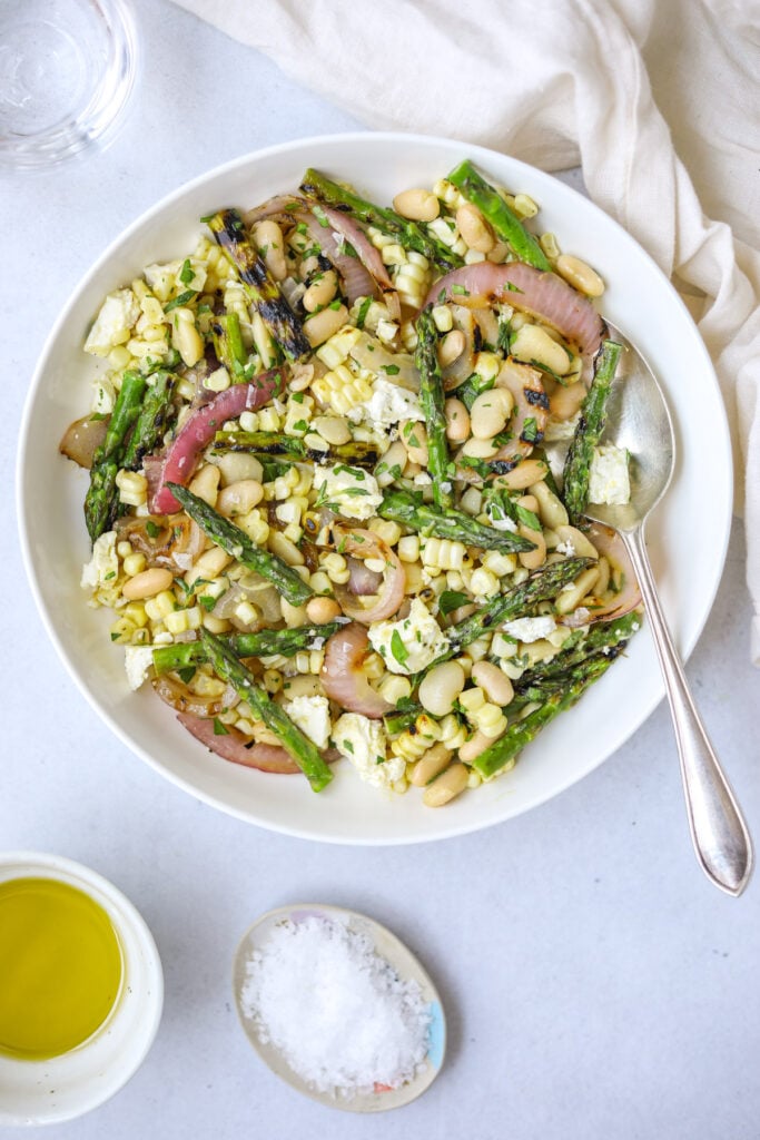 a white bowl filled with grilled vegetable salad with white beans, get feta cheese, asparagus, red onion and silver spoon on the side