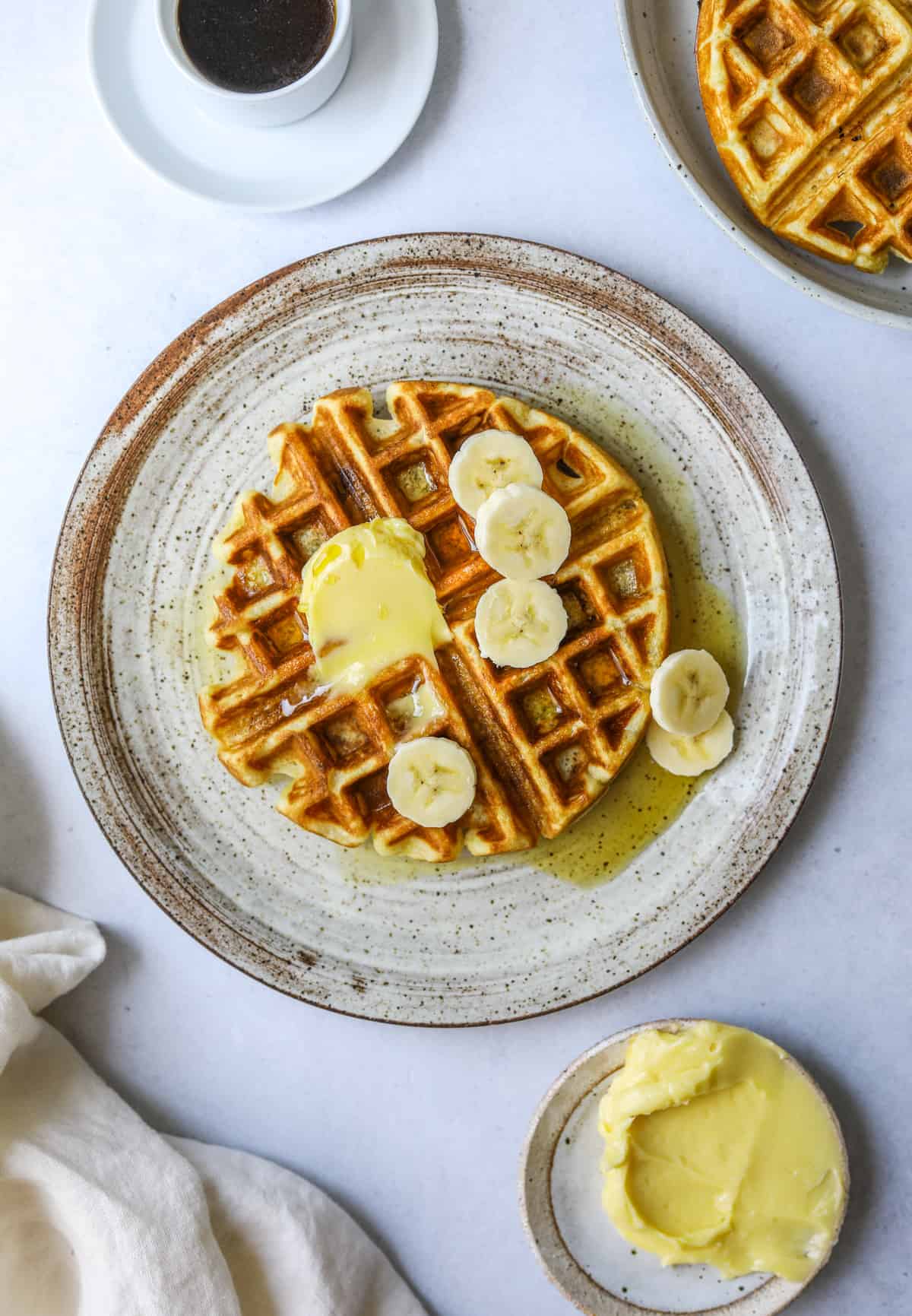a ceramic plate with a banana waffle topped with fresh bananas, maple syrup and butter