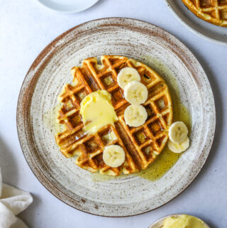 a ceramic plate with a waffle topped with banana slices, a cup of coffee and a dish of butter