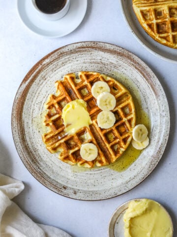 a ceramic plate with a waffle topped with banana slices, a cup of coffee and a dish of butter