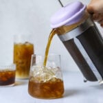 a french press pouring cold brew iced coffee into a clear glass of ice