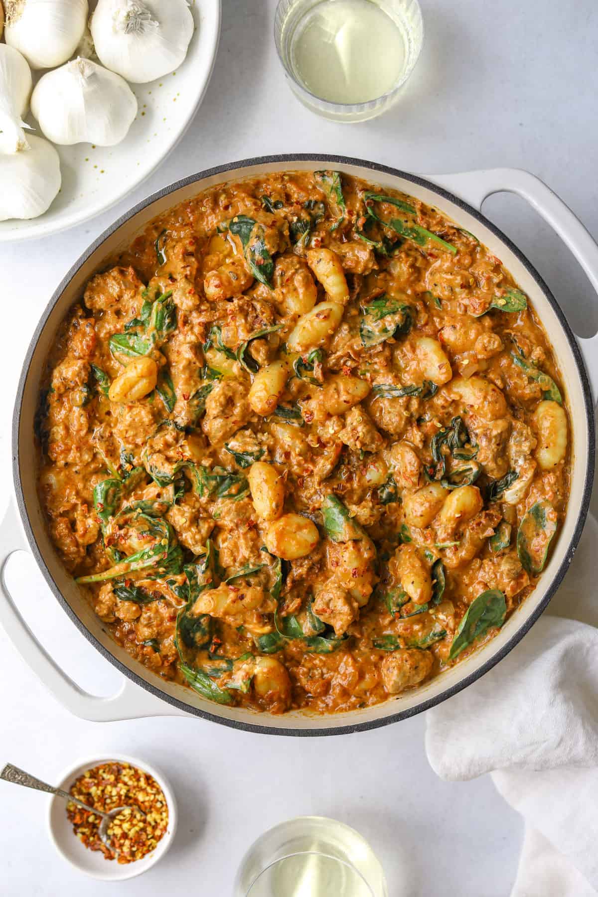 a dutch oven skillet filled with sun-dried tomato gnocchi with a bowl of garlic, a glass of white wine on a blue background