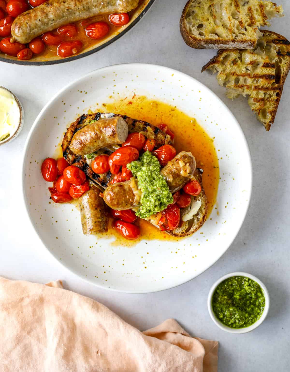 a white bowl of sausages with tomatoes and grilled bread
