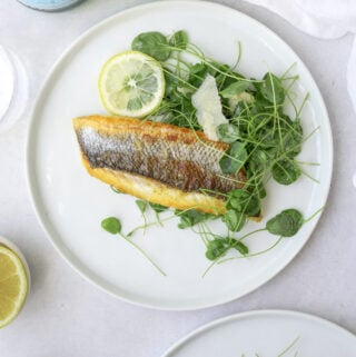 a white plate on a light blue background with branzino filets, watercress and lemon slices