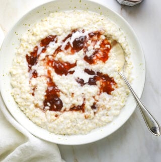 a ceramic bowl filled with brown rice porridge topped with strawberry jam with a white tea towel on a stone background