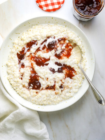 a ceramic bowl filled with brown rice porridge topped with strawberry jam with a white tea towel on a stone background