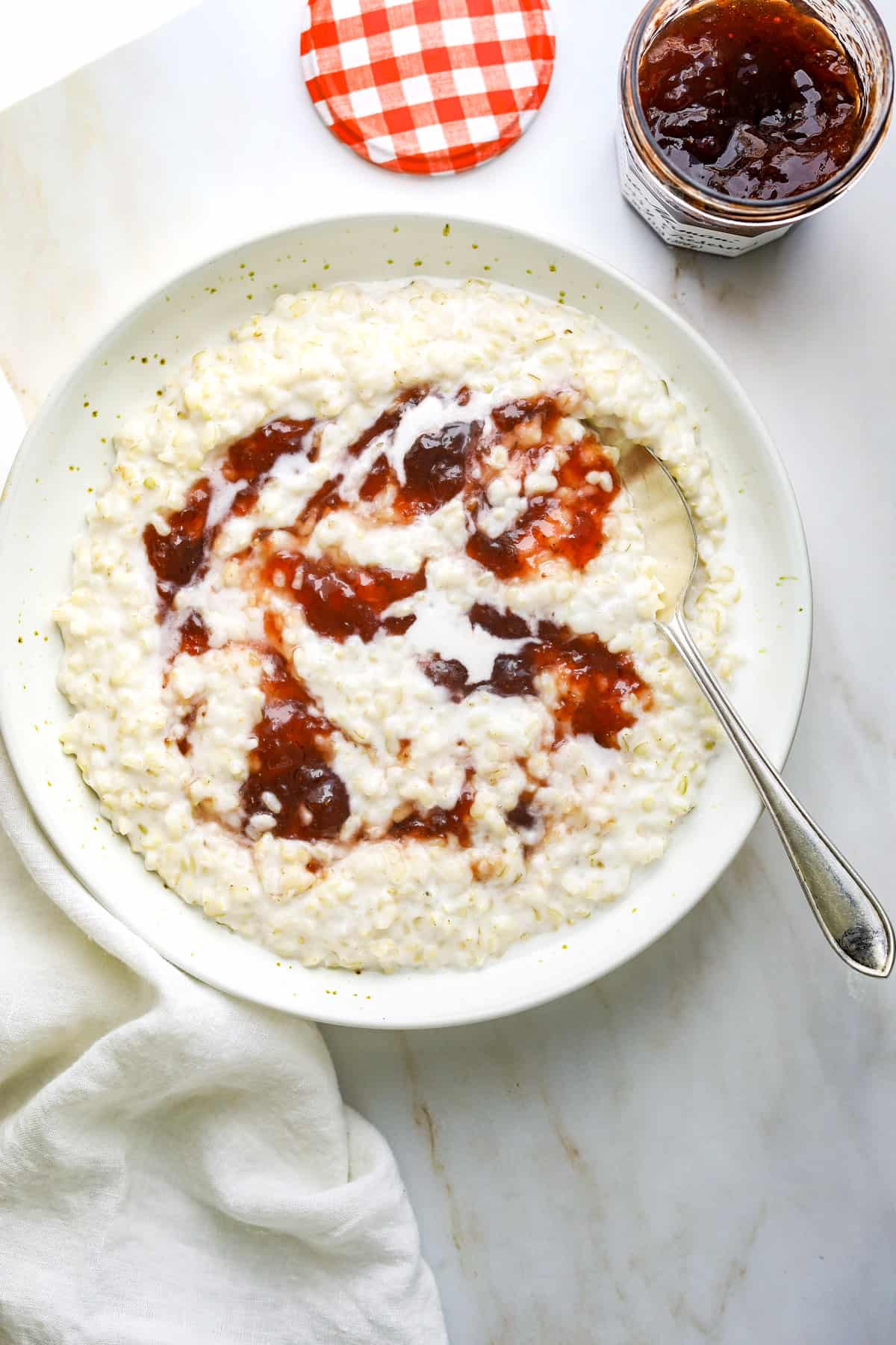 a ceramic bowl filled with brown rice porridge topped with strawberry jam with a white tea towel on a stone background