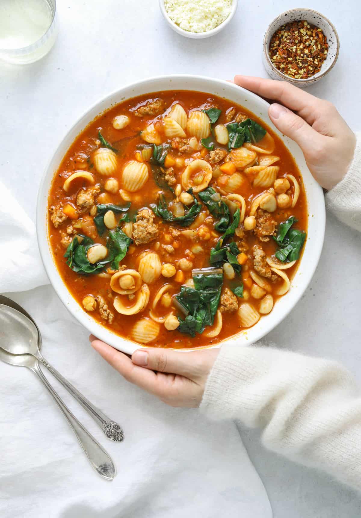 a bowl of soup with two hands holding it and spoons on the side