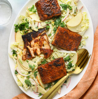 a white oval platter with 4 filets of blackened salmon on top of cabbage slaw on a grey background and gold serving utensils