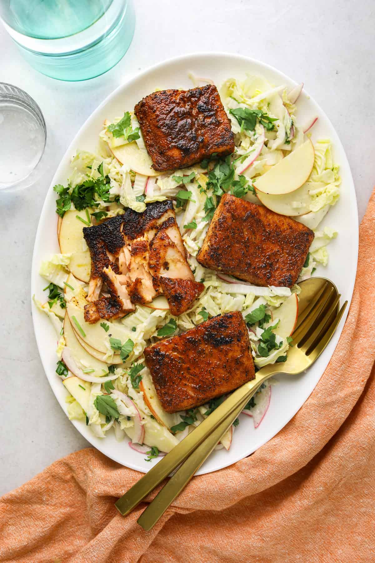 a white oval platter with 4 filets of blackened salmon on top of cabbage slaw on a grey background and gold serving utensils