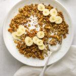 a bowl of banana bread granola with sliced bananas and a silver spoon with a white tea towel