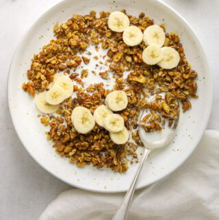 a bowl of banana bread granola with sliced bananas and a silver spoon with a white tea towel