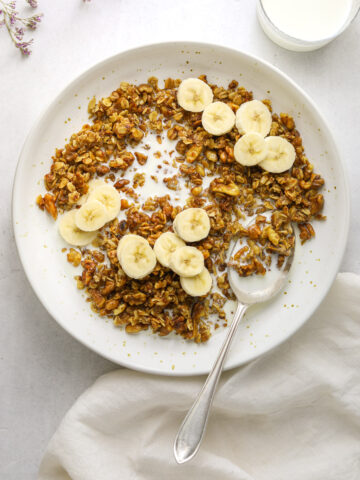 a bowl of banana bread granola with sliced bananas and a silver spoon with a white tea towel