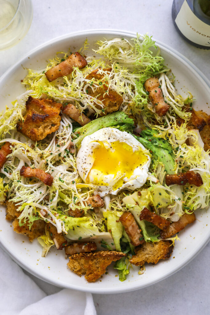 a close up photo of frisée salad in a white bowl with a poached egg with runny yolk, bacon and croutons