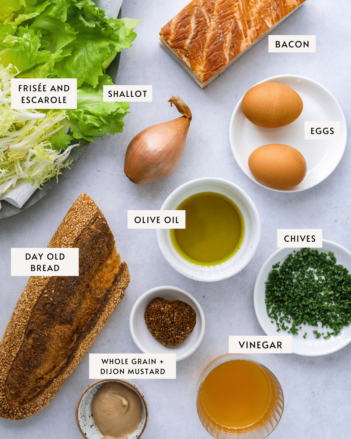 recipe ingredients on a blue background individually portioned and labeled: lettuce, eggs, baguette, bacon, chives, oil, vinegar, mustard.