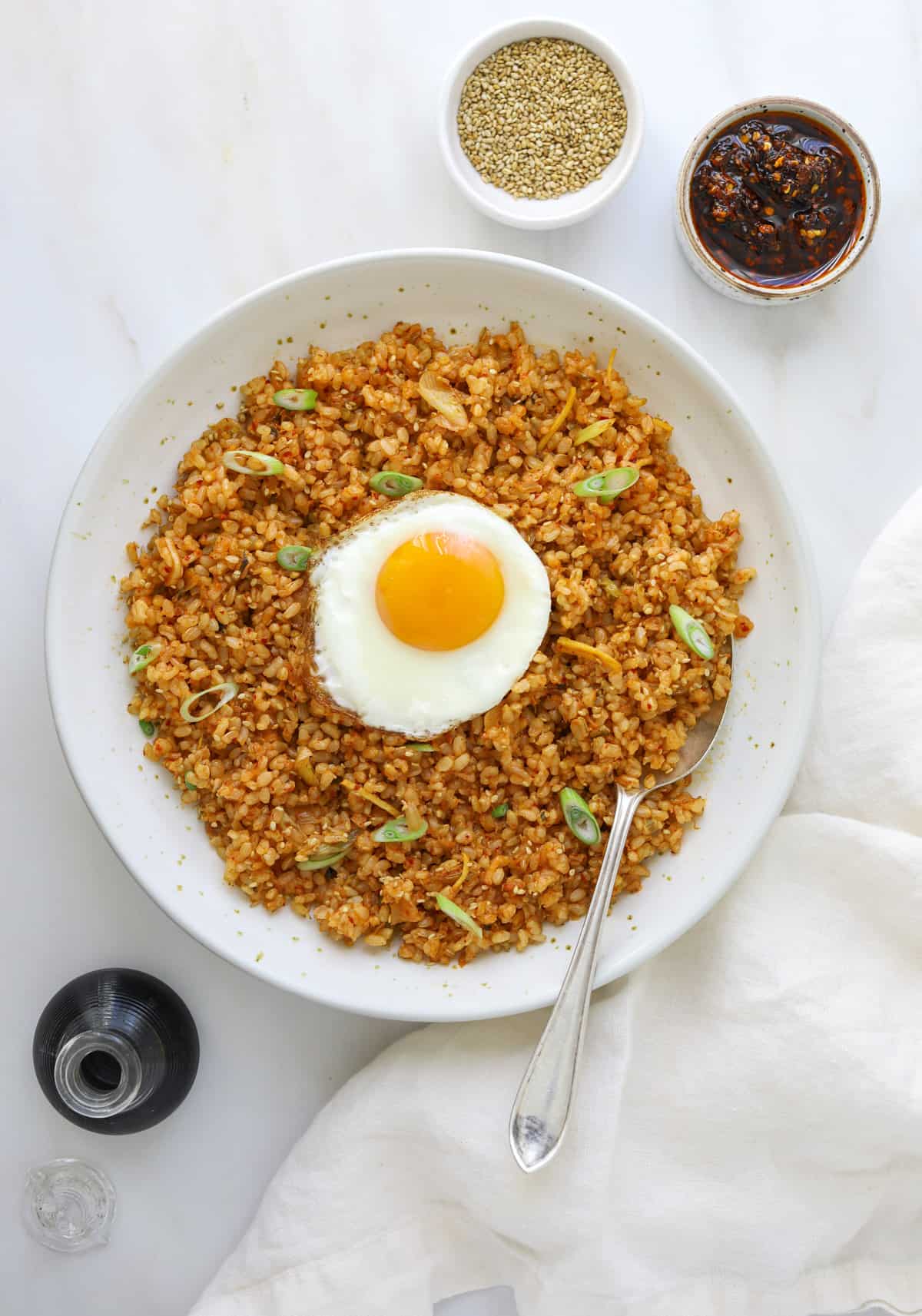 a ceramic bowl filled with fried rice, topped with a sunny side up egg on a marble background with a silver spoon and white napkin