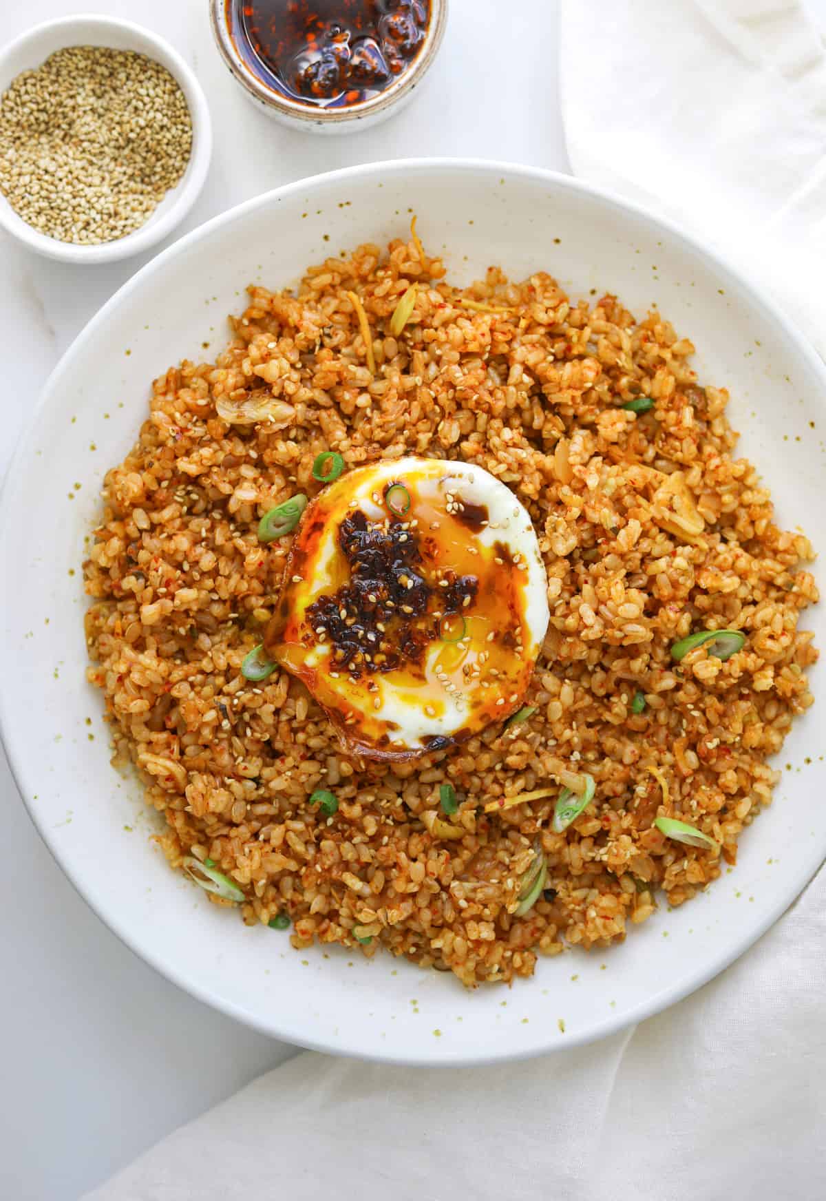 a bowl of kimchi fried rice topped with a runny egg, chili oil and thinly sliced green onions with a bowl of toasted sesame seeds on the side