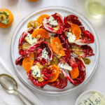 a radicchio and persimmon salad on a white plate with blue cheese a white napkin and a glass of wine