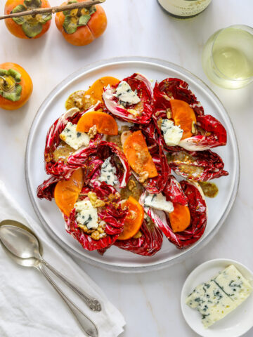 a radicchio and persimmon salad on a white plate with blue cheese a white napkin and a glass of wine
