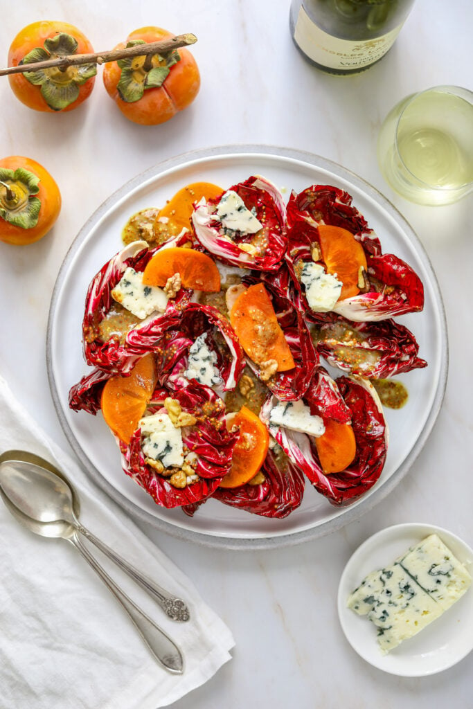 a radicchio and persimmon salad on a white plate with blue cheese a white napkin and a glass of wine