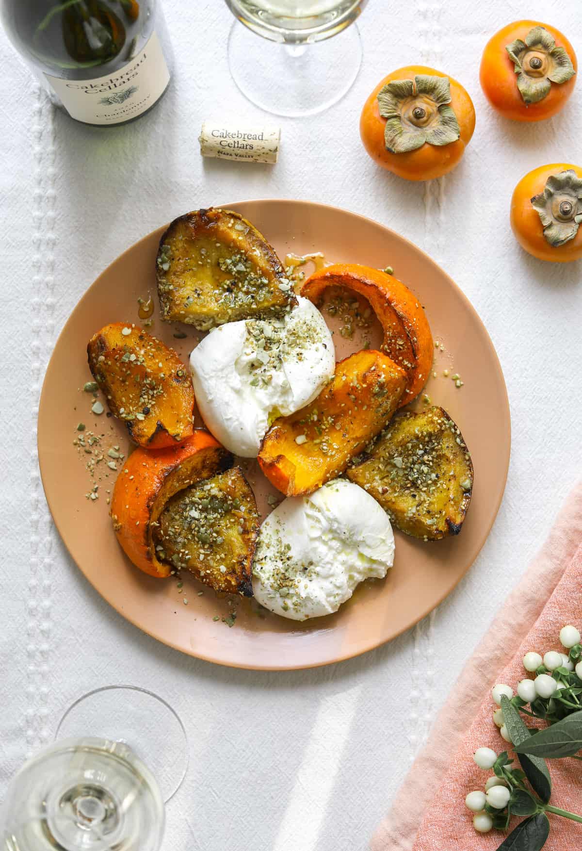 a place of roasted squash and burrata on a linen table cloth with linen napkins, persimmons and flowers with bottle of wine