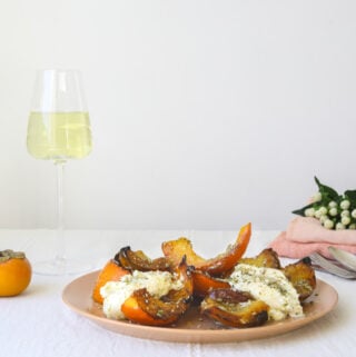 a plate of roasted squash on a cloth lined table with two glasses of white wine, pink linen napkins and flowers