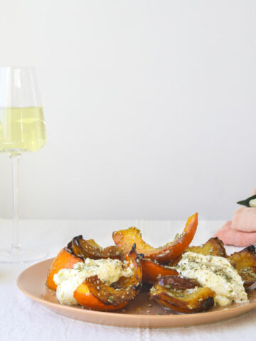 a plate of roasted squash on a cloth lined table with two glasses of white wine, pink linen napkins and flowers