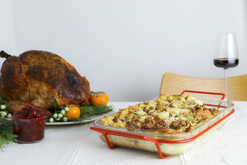 a table setting with a white table cloth, a whole roasted turkey, a glass off red wine and a baking dish with savory bread pudding