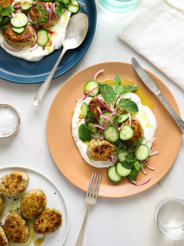 a marble table top with a peach colored plate filled with yogurt and chicken koftas, herbs, cucumbers and red onion