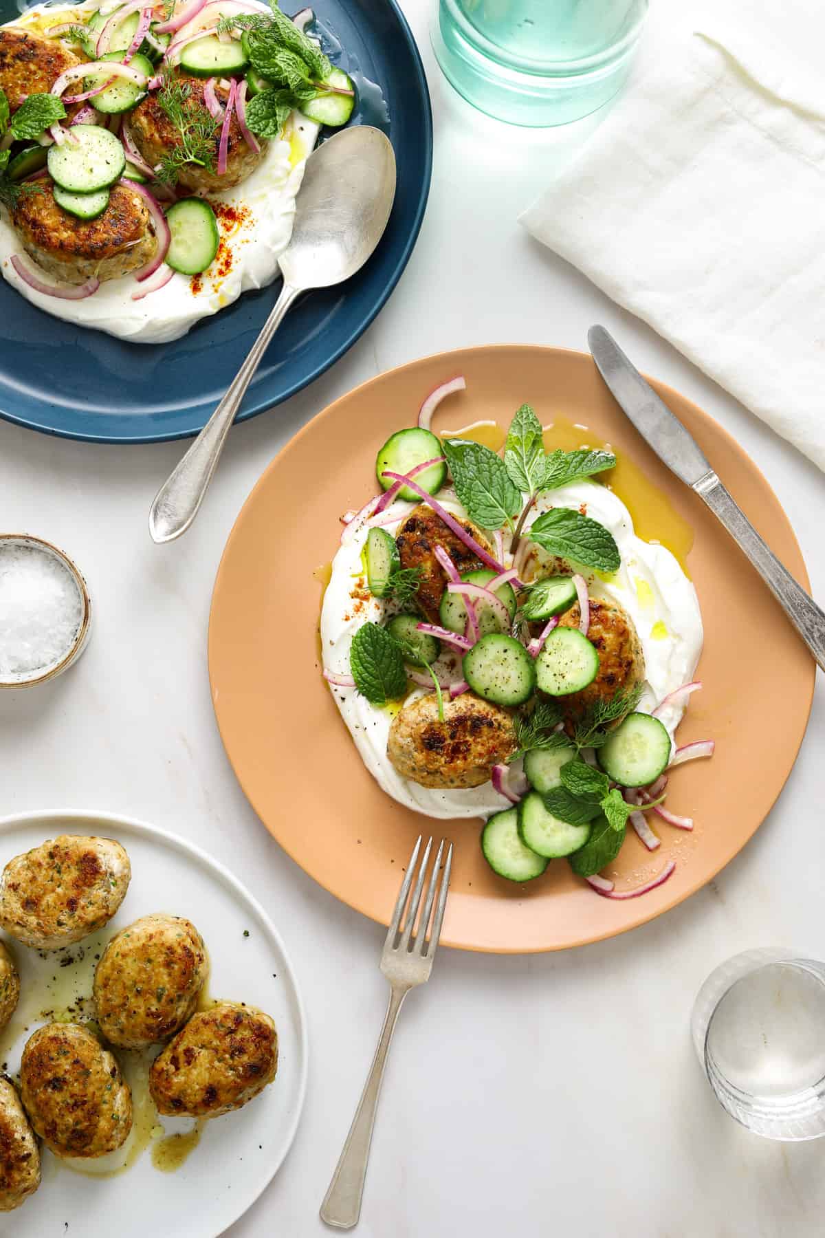 a marble table top with a peach colored plate filled with yogurt and chicken koftas, herbs, cucumbers and red onion