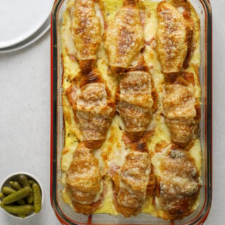 9 croque monsieur croissants in a clear glass baking dish with red handles. two side dishes with pickles and dijon mustard