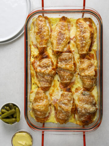 9 croque monsieur croissants in a clear glass baking dish with red handles. two side dishes with pickles and dijon mustard