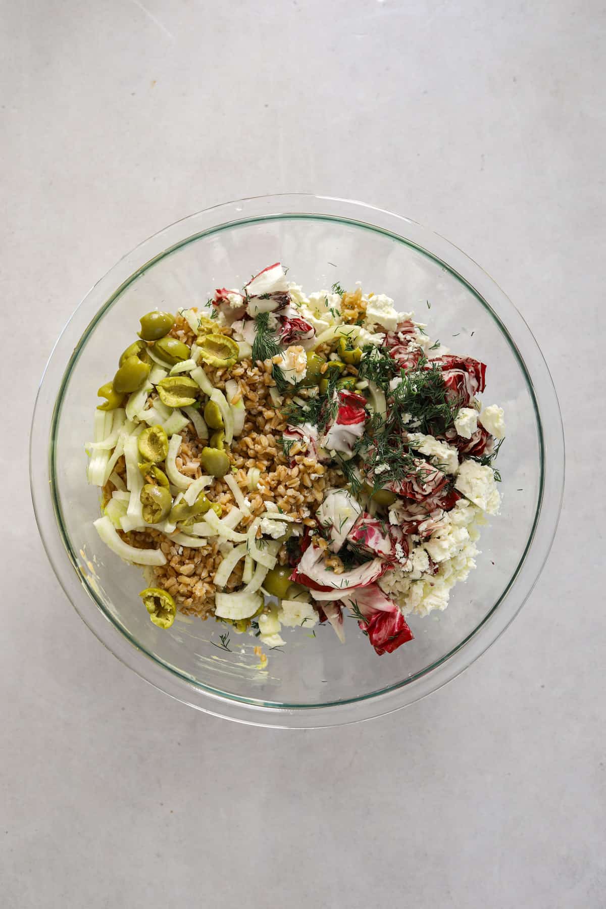 a glass mixing bowl filled with farro salad ingredients on a gray background
