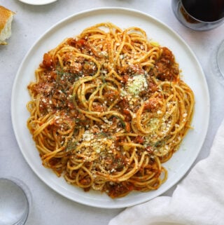 a bowl of spaghetti on a white plate on a light blue background with a glass of red wine, a glass of water and a baguette