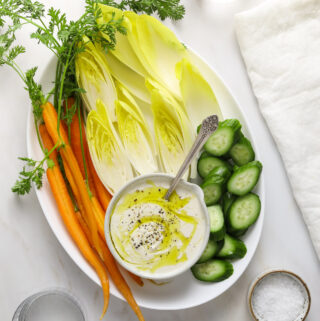 a white platter with carrots, cucumber, endive and small bowl of tahini labneh dip