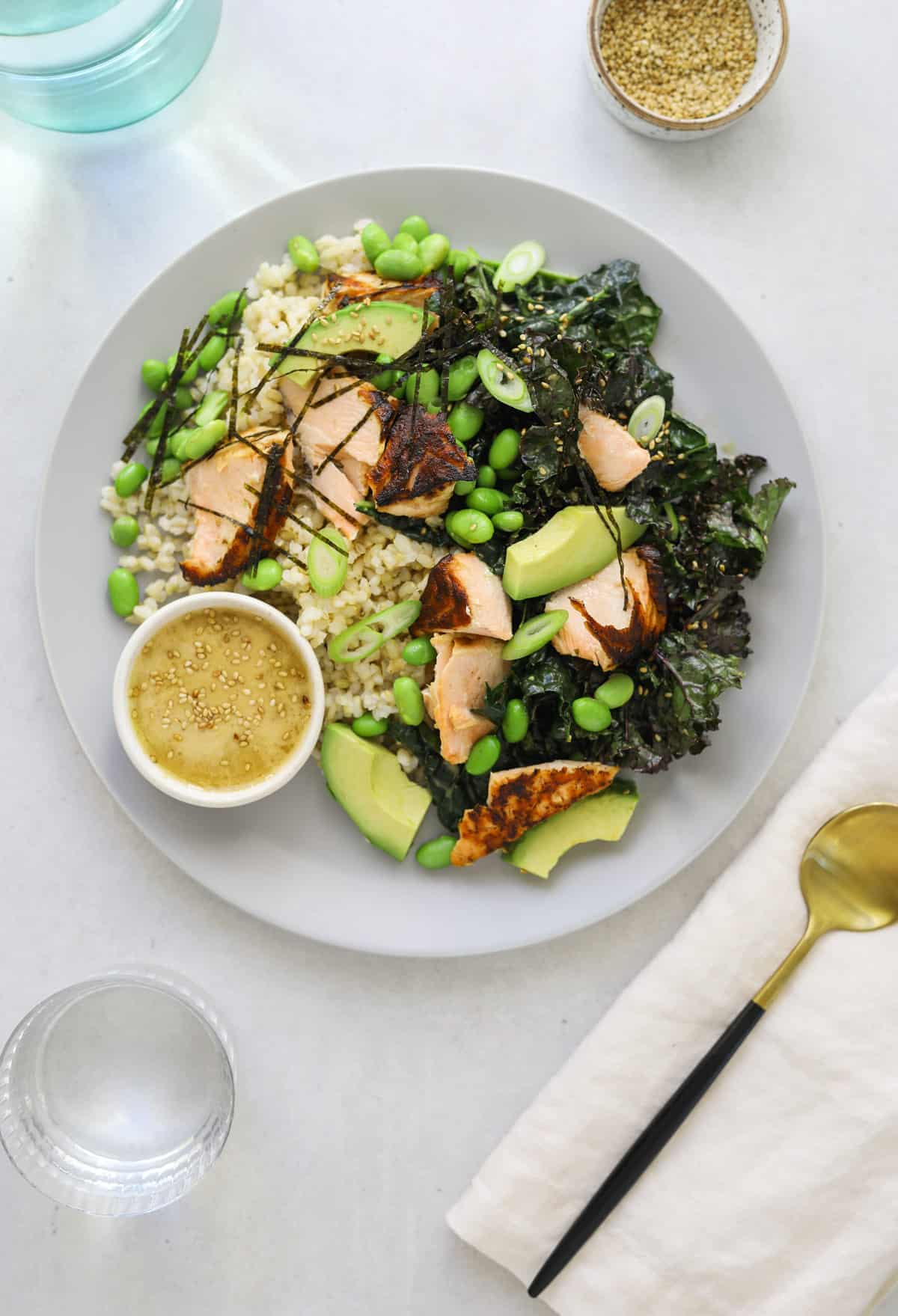 a light grey plate filled with brown rice, kale, avocado and salmon salad on a blue background with a gold and black spoon and white linen napkin